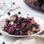 Blueberry dessert cobbler with mixed berries on a white plate surrounded by fresh blueberries and a beige napkin, served as a delicious sweet treat.