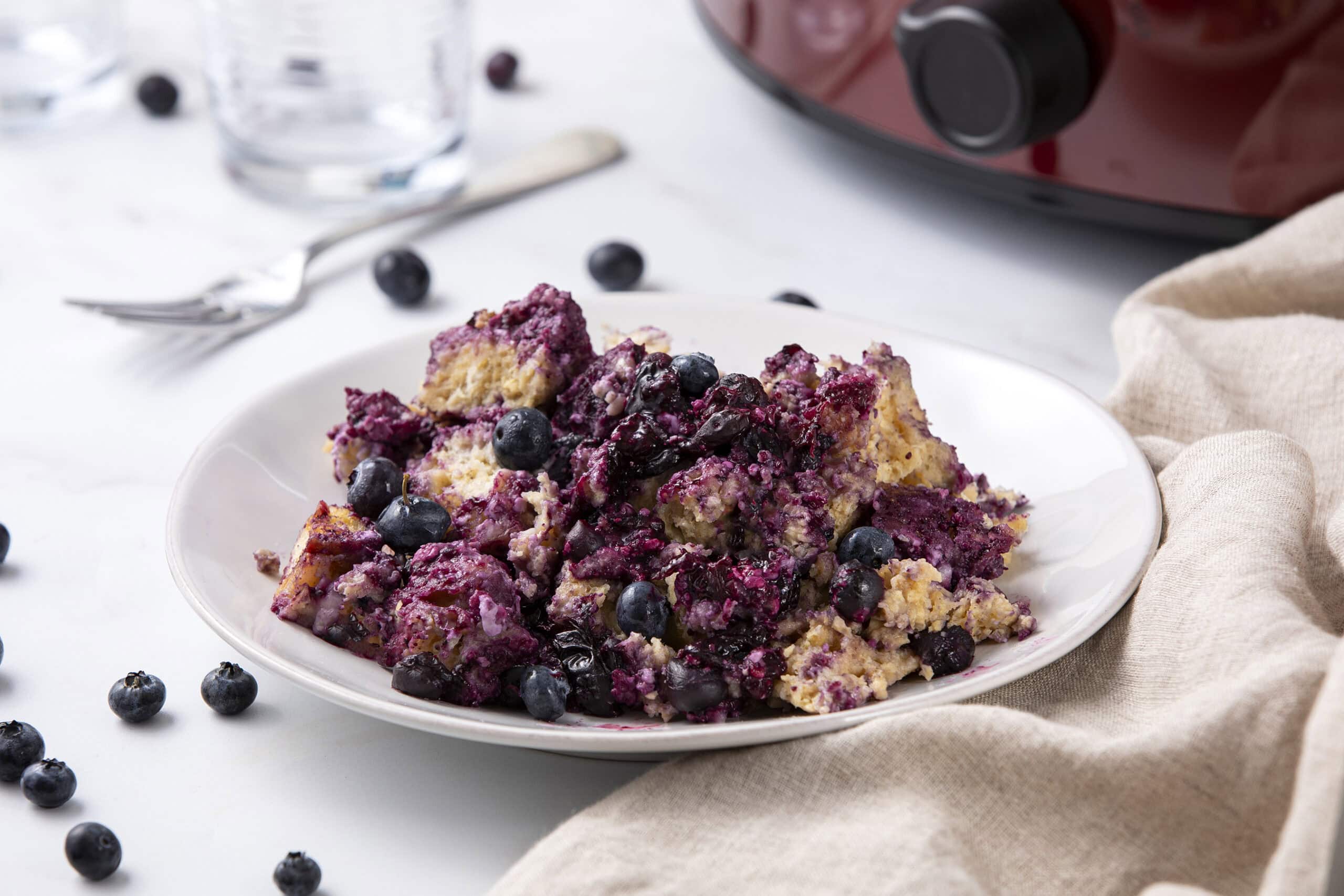 Blueberry dessert cobbler with mixed berries on a white plate surrounded by fresh blueberries and a beige napkin, served as a delicious sweet treat.