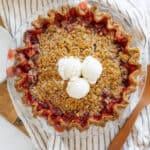 Sweet cherry pie with crumbly topping, served with vanilla ice cream scoops on top, in a clear glass baking dish on a wooden board.