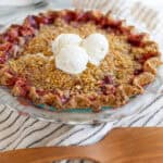 Sweet cherry crumble pie topped with vanilla ice cream, served in a glass pie dish on a striped cloth. Perfect for dessert, featuring a crispy crumble topping and juicy cherry filling.