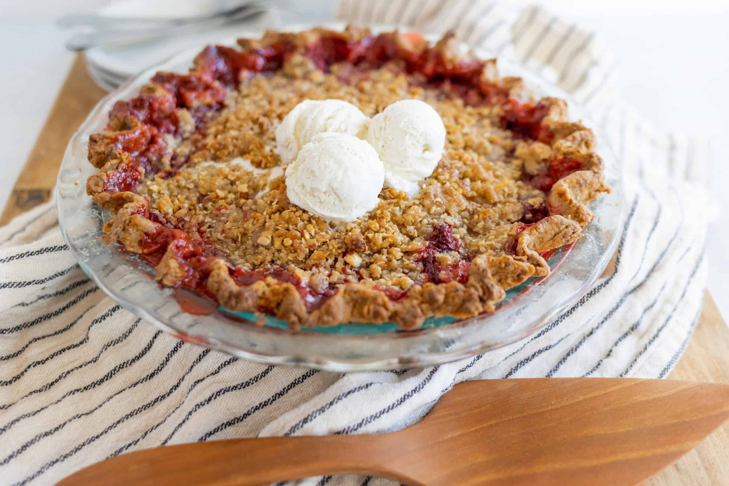 Sweet cherry crumble pie topped with vanilla ice cream, served in a glass pie dish on a striped cloth. Perfect for dessert, featuring a crispy crumble topping and juicy cherry filling.