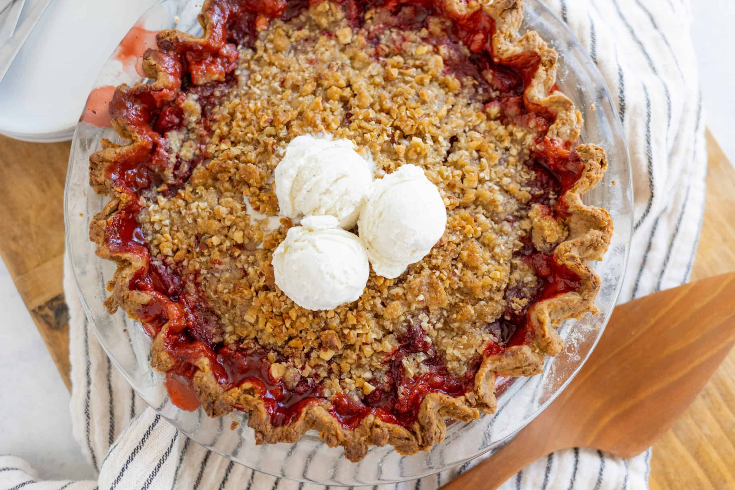Fresh homemade cherry crumble pie with a flaky crust, juicy cherry filling, crunchy oat topping, and scoops of vanilla ice cream on top, served in a clear glass pie dish.