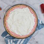Fresh strawberry pie with a graham cracker crust, topped with whipped cream, on a marble surface with strawberries and a red spatula.