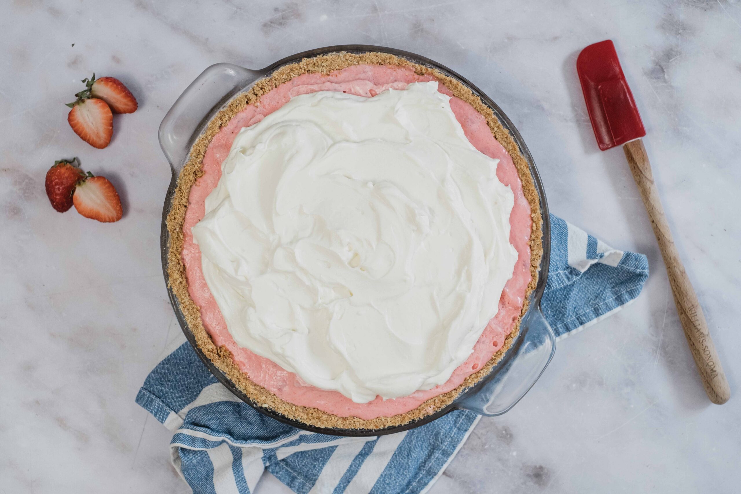 Fresh strawberry pie with a graham cracker crust, topped with whipped cream, on a marble surface with strawberries and a red spatula.
