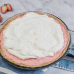 Fresh strawberry Jello pie with whipped topping in a clear glass dish on a marble surface, ready for baking or serving.