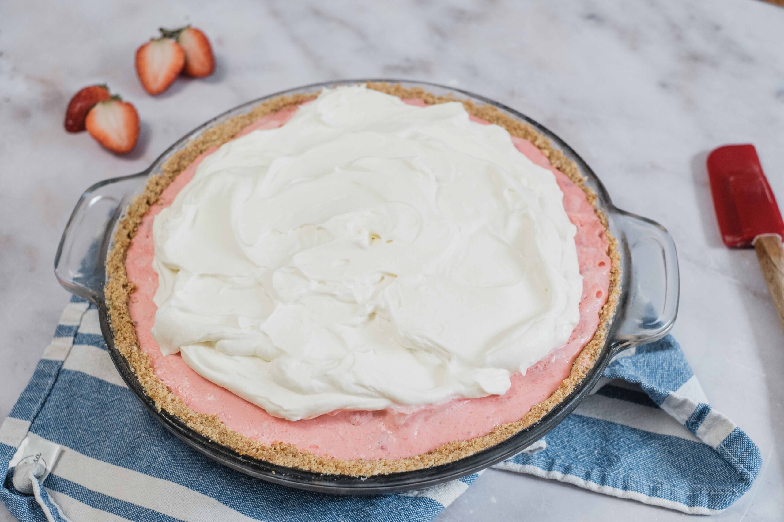 Fresh strawberry Jello pie with whipped topping in a clear glass dish on a marble surface, ready for baking or serving.