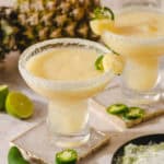 Refreshing pineapple margarita cocktails garnished with lime and jalapeño slices, served in salted rim glasses, perfect for summer parties and tropical vacations.