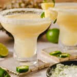 Freshly made lime margaritas in elegant glasses with salted rims, garnished with lime wedges, served on a table with fresh lime and jalapeño peppers for a spicy twist; perfect for summer gatherings or celebrations.