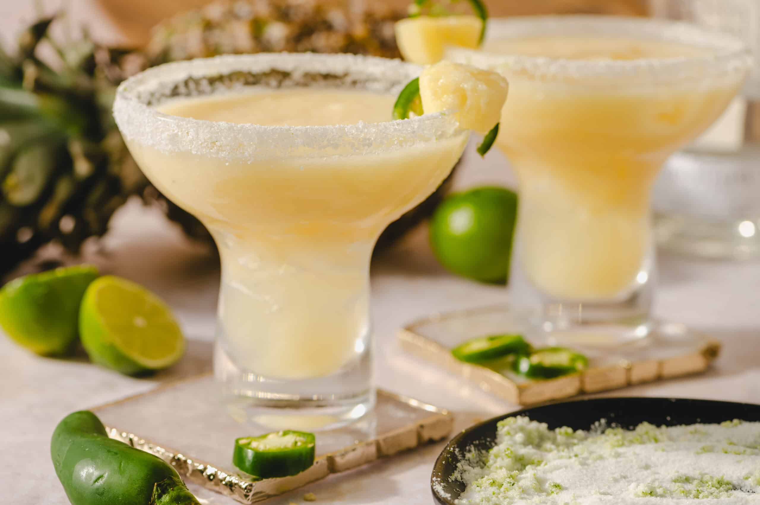 Freshly made lime margaritas in elegant glasses with salted rims, garnished with lime wedges, served on a table with fresh lime and jalapeño peppers for a spicy twist; perfect for summer gatherings or celebrations.
