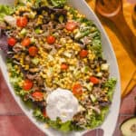 Fresh taco salad with ground beef, cherry tomatoes, avocado, corn, shredded cheese, and lettuce on a white platter, served with a dollop of sour cream, on a rustic red tile surface.