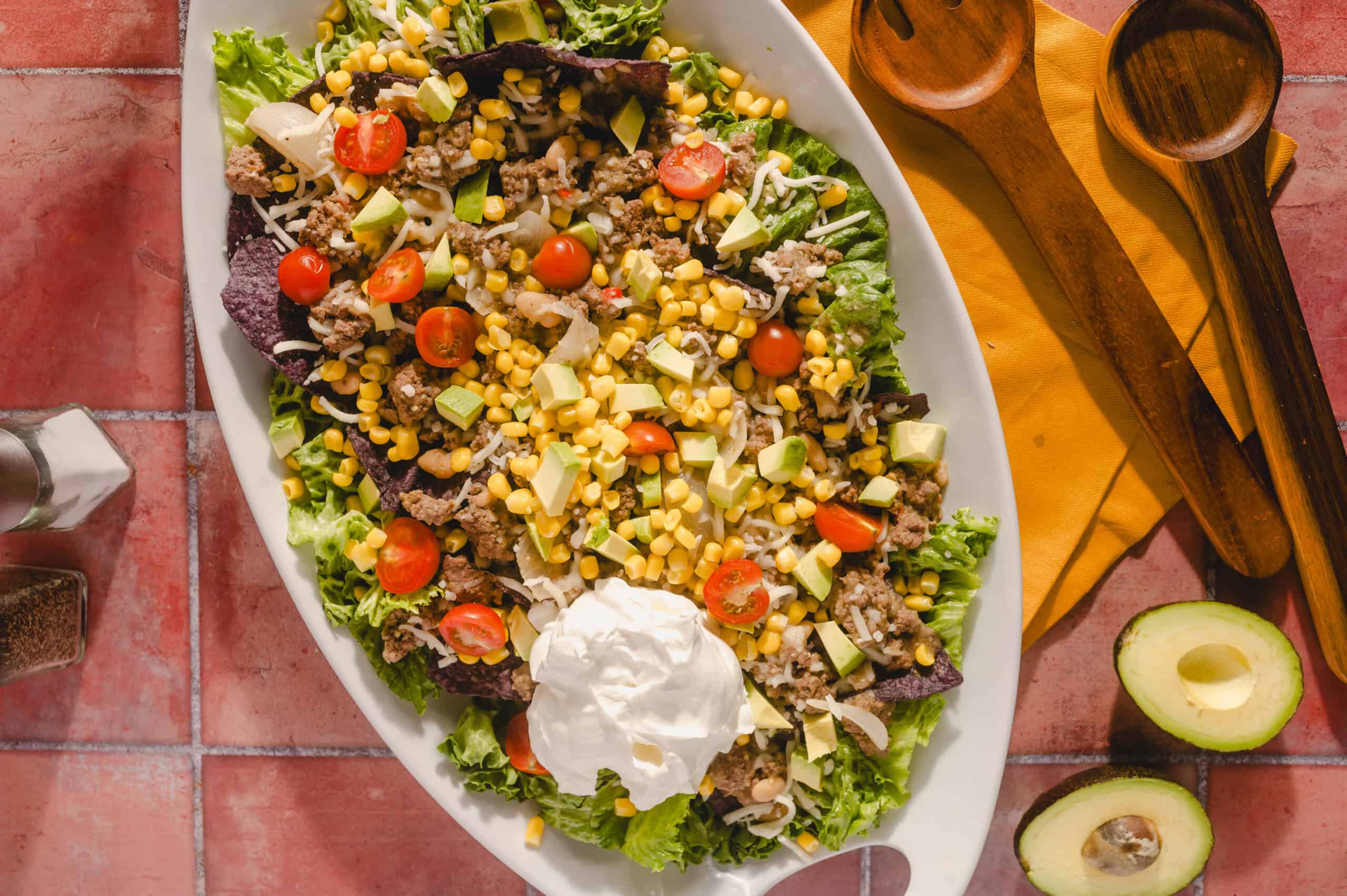 Fresh taco salad with ground beef, cherry tomatoes, avocado, corn, shredded cheese, and lettuce on a white platter, served with a dollop of sour cream, on a rustic red tile surface.