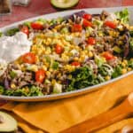 Fresh taco salad with ground beef, cherry tomatoes, avocado, corn, shredded cheese, and sour cream on a white platter. Perfect for a healthy, delicious meal or party dish.