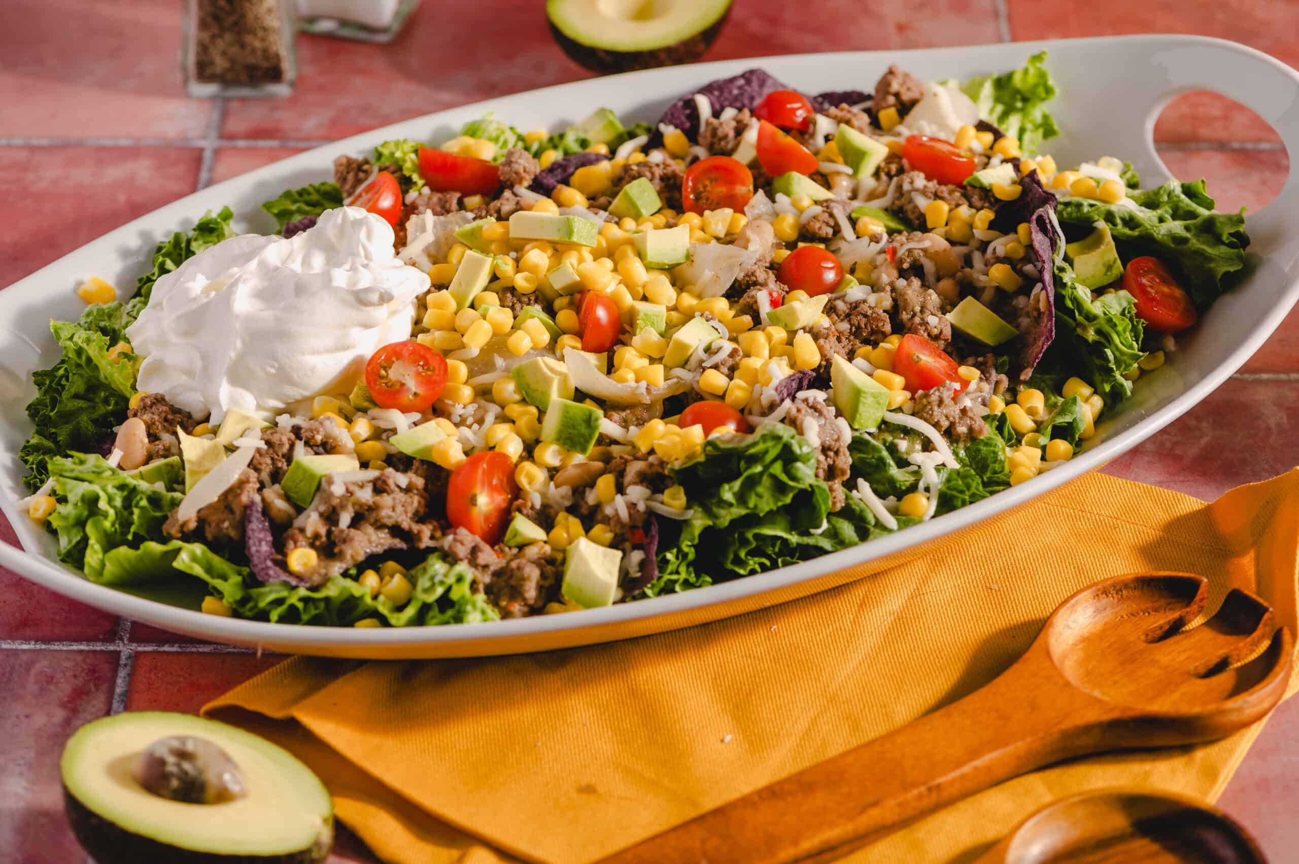 Fresh taco salad with ground beef, cherry tomatoes, avocado, corn, shredded cheese, and sour cream on a white platter. Perfect for a healthy, delicious meal or party dish.
