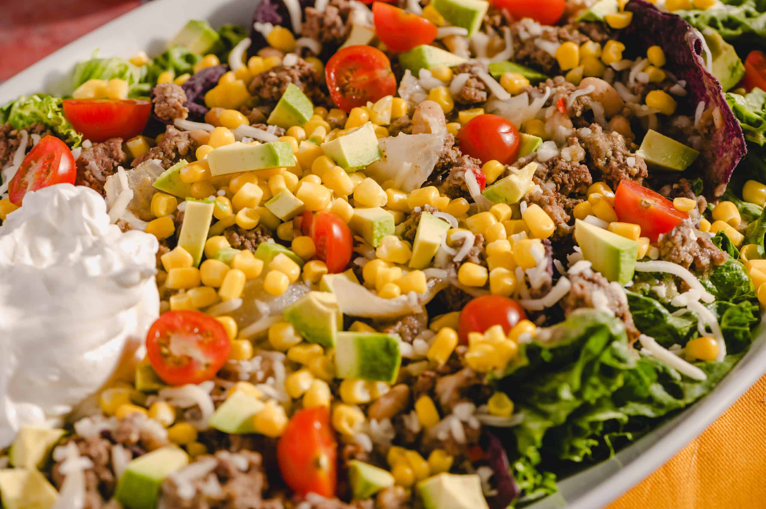 Fresh taco salad with ground beef, cherry tomatoes, avocado, sweet corn, shredded cheese, and lettuce on a white platter. Perfect for healthy Mexican-inspired meal ideas.