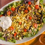 Fresh taco salad with ground beef, cherry tomatoes, avocado, corn, shredded cheese, lettuce, and sour cream on a white serving platter. Perfect for a flavorful, healthy meal or party appetizer.