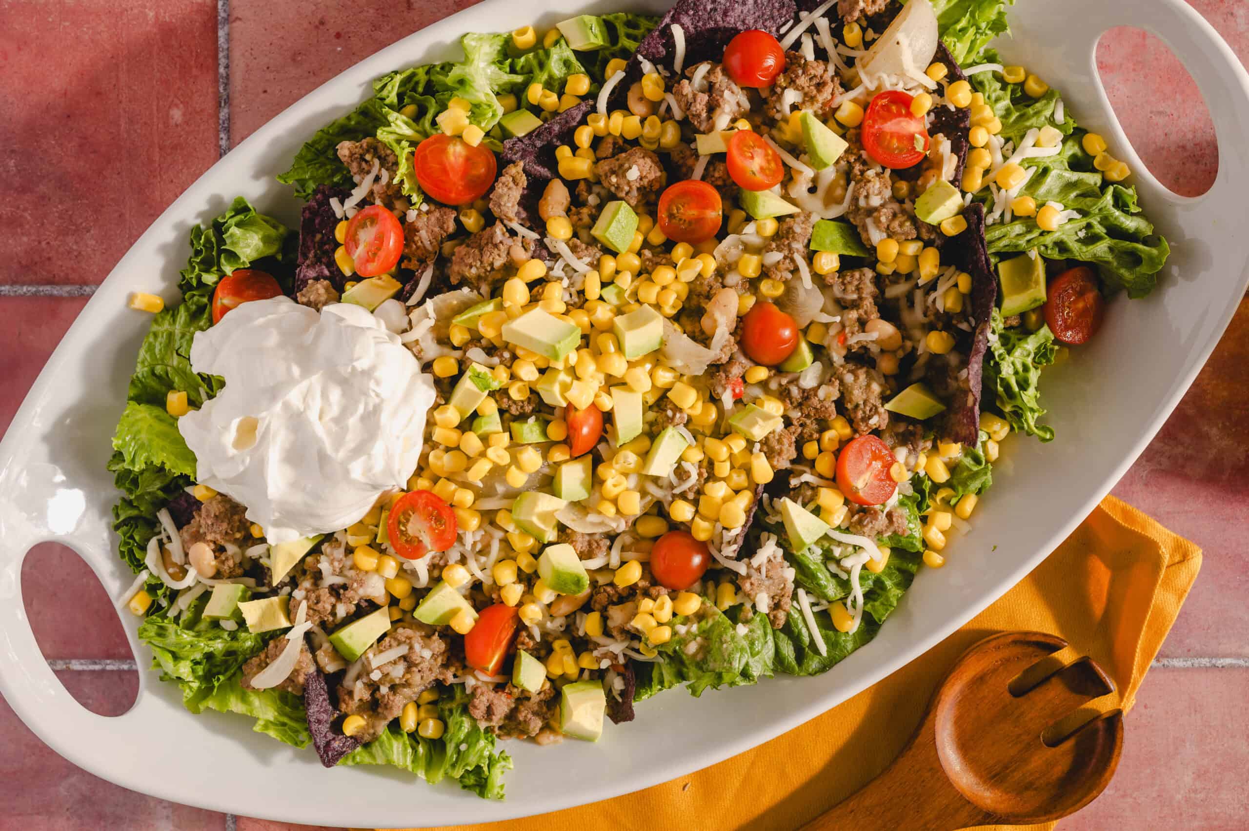 Fresh taco salad with ground beef, cherry tomatoes, avocado, corn, shredded cheese, lettuce, and sour cream on a white serving platter. Perfect for a flavorful, healthy meal or party appetizer.