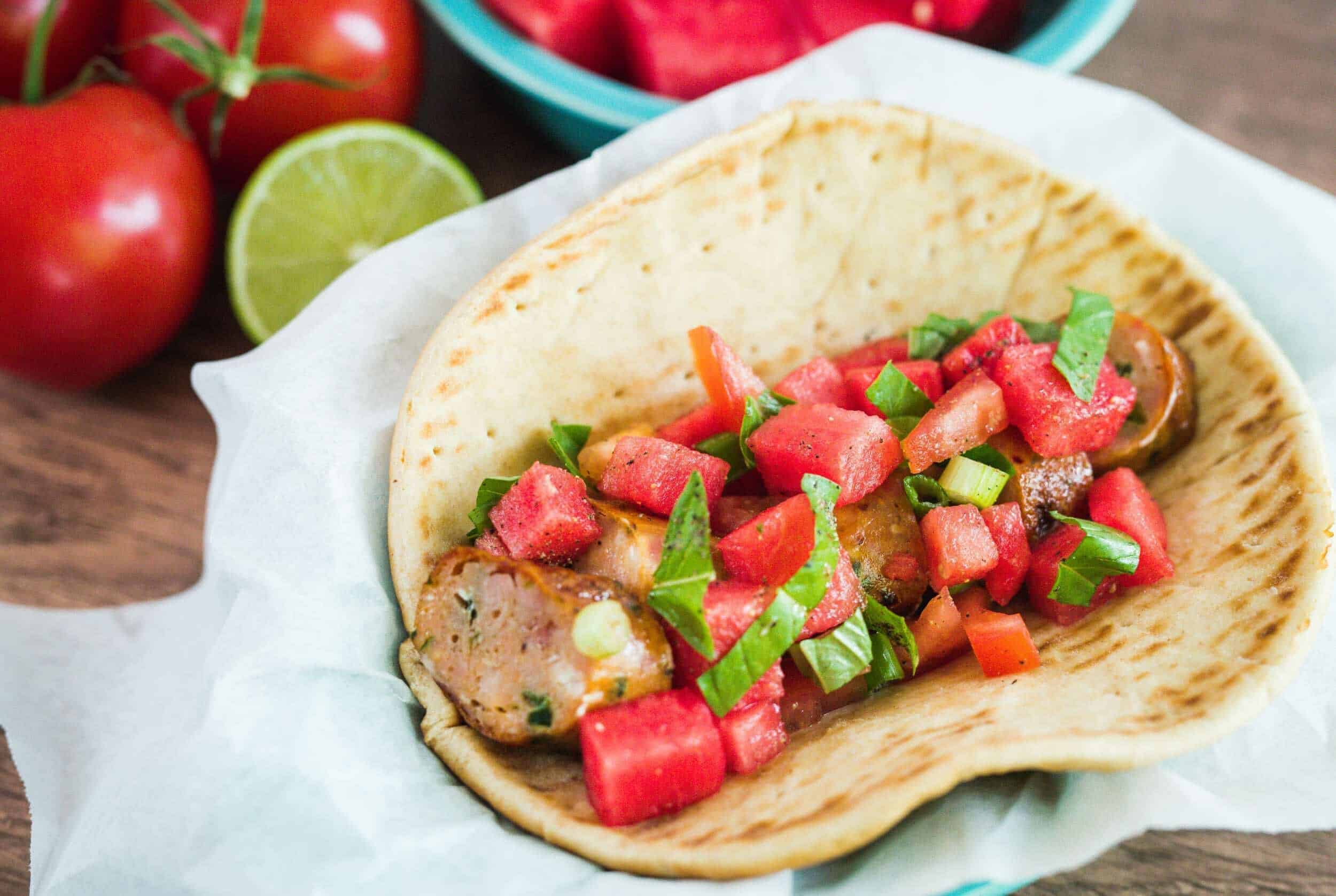 Fresh grilled sausage taco with diced tomatoes and green onions served on a soft flour tortilla, with fresh tomatoes and lime in the background for a tasty Mexican-inspired dish.