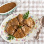 Tender beef stew with rich gravy served on a floral plate garnished with parsley, accompanied by a side of hearty brown sauce or gravy in a small pitcher, on a checkered tablecloth.