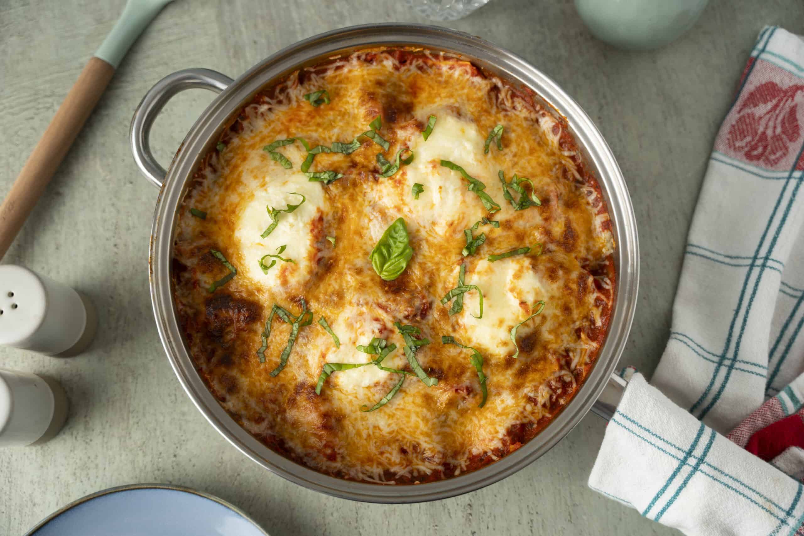 Creamy baked lasagna in a stainless steel dish topped with melted cheese, fresh basil, and chopped herbs, served on a rustic kitchen countertop for a delicious Italian home-cooked meal.