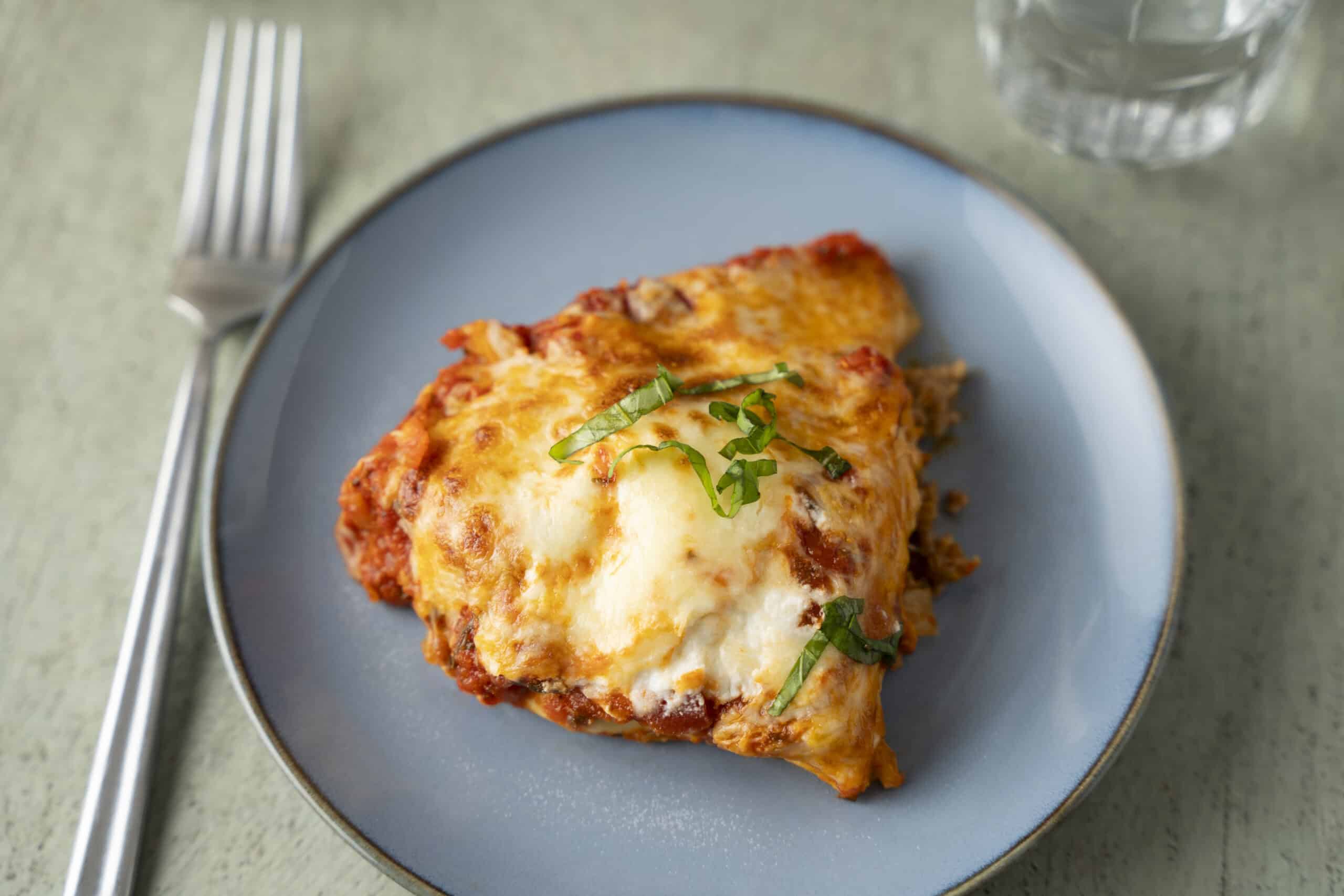 Tender baked lasagna with melted cheese and fresh basil garnish on a blue plate. Perfect comfort food for dinner, featuring rich tomato sauce, gooey cheese, and perfectly layered pasta.
