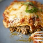 Hearty homemade lasagna with layers of seasoned ground beef, tomato sauce, ricotta, and melted cheese topped with fresh basil, served on a dark plate.