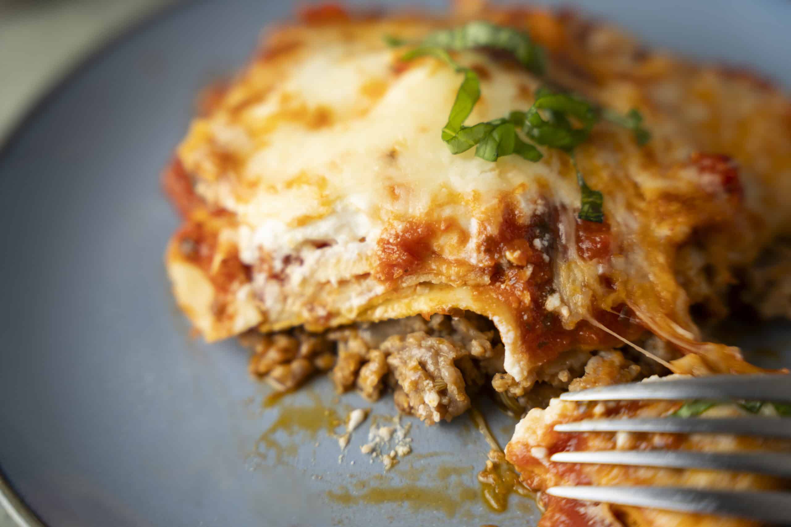Hearty homemade lasagna with layers of seasoned ground beef, tomato sauce, ricotta, and melted cheese topped with fresh basil, served on a dark plate.