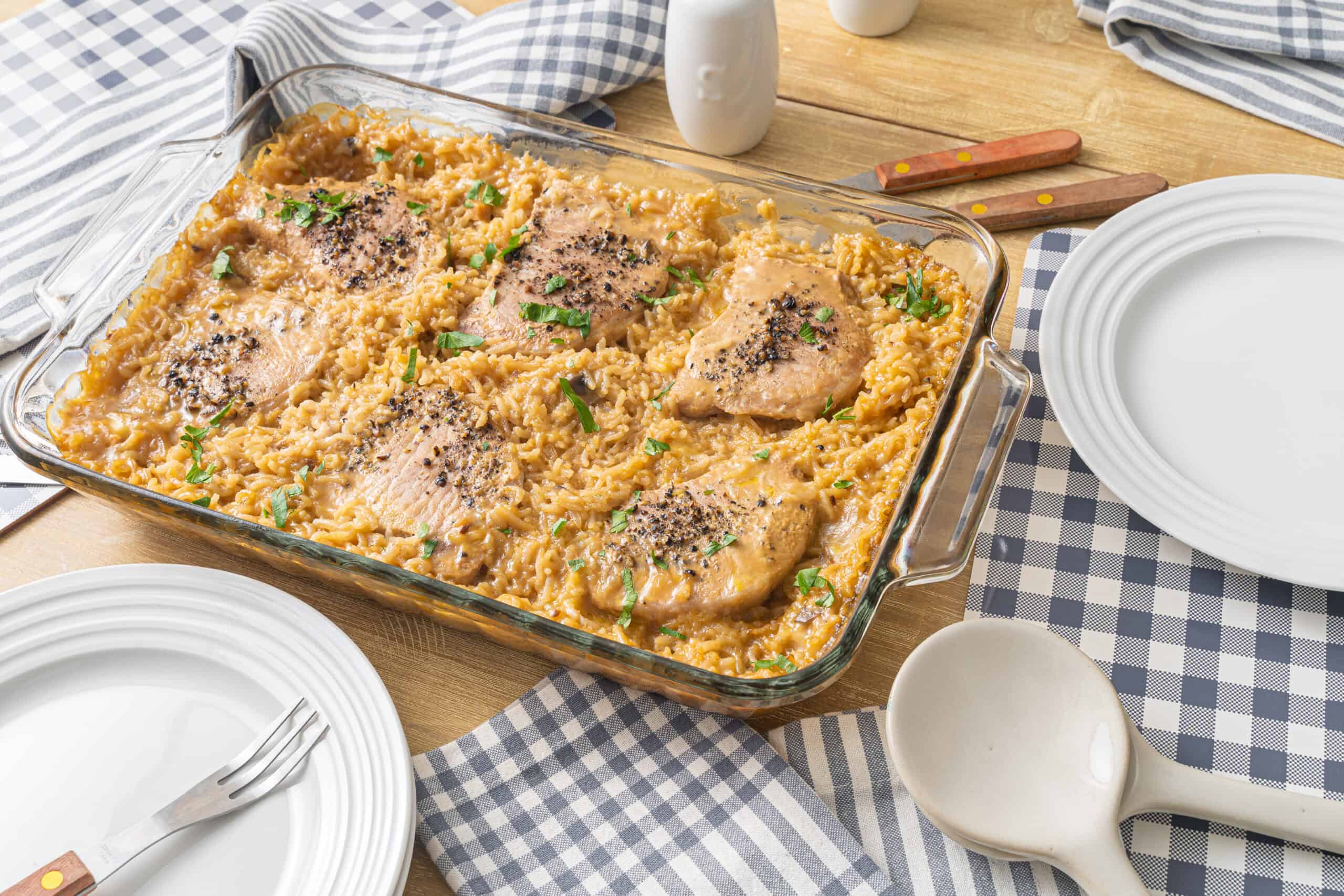 Creamy chicken and rice casserole baked with melted cheese, topped with herbs and black pepper, served in a glass dish on a wooden table with white plates and striped napkins.