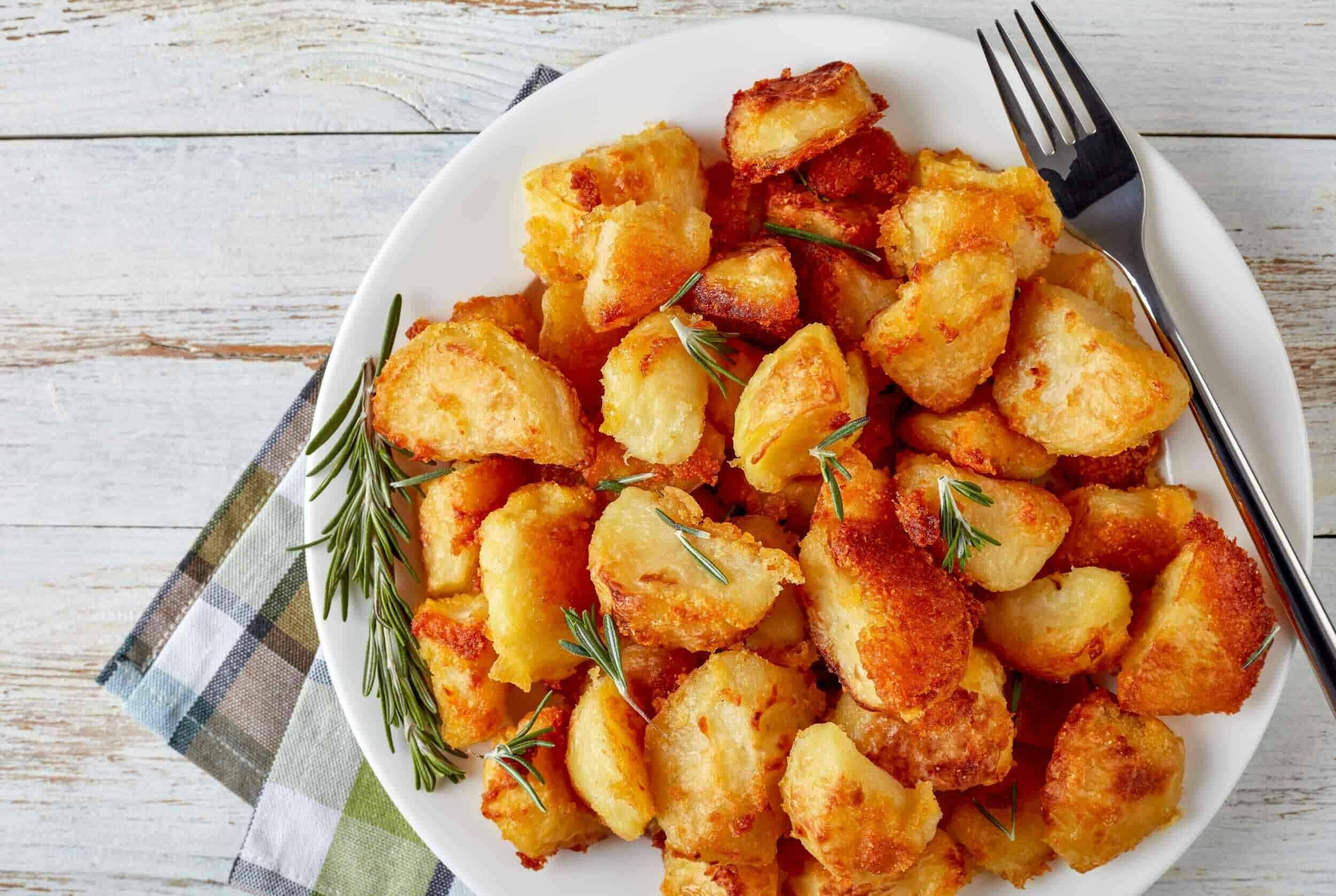 Crispy golden roasted potatoes garnished with fresh rosemary on a white plate. Perfect side dish for breakfast, lunch, or dinner, served on a rustic wooden table with a fork.