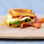 Savory breakfast sandwich with crispy bacon, fried egg, fresh avocado slices, and toasted bread on wooden cutting board. Perfect for morning meal or brunch.