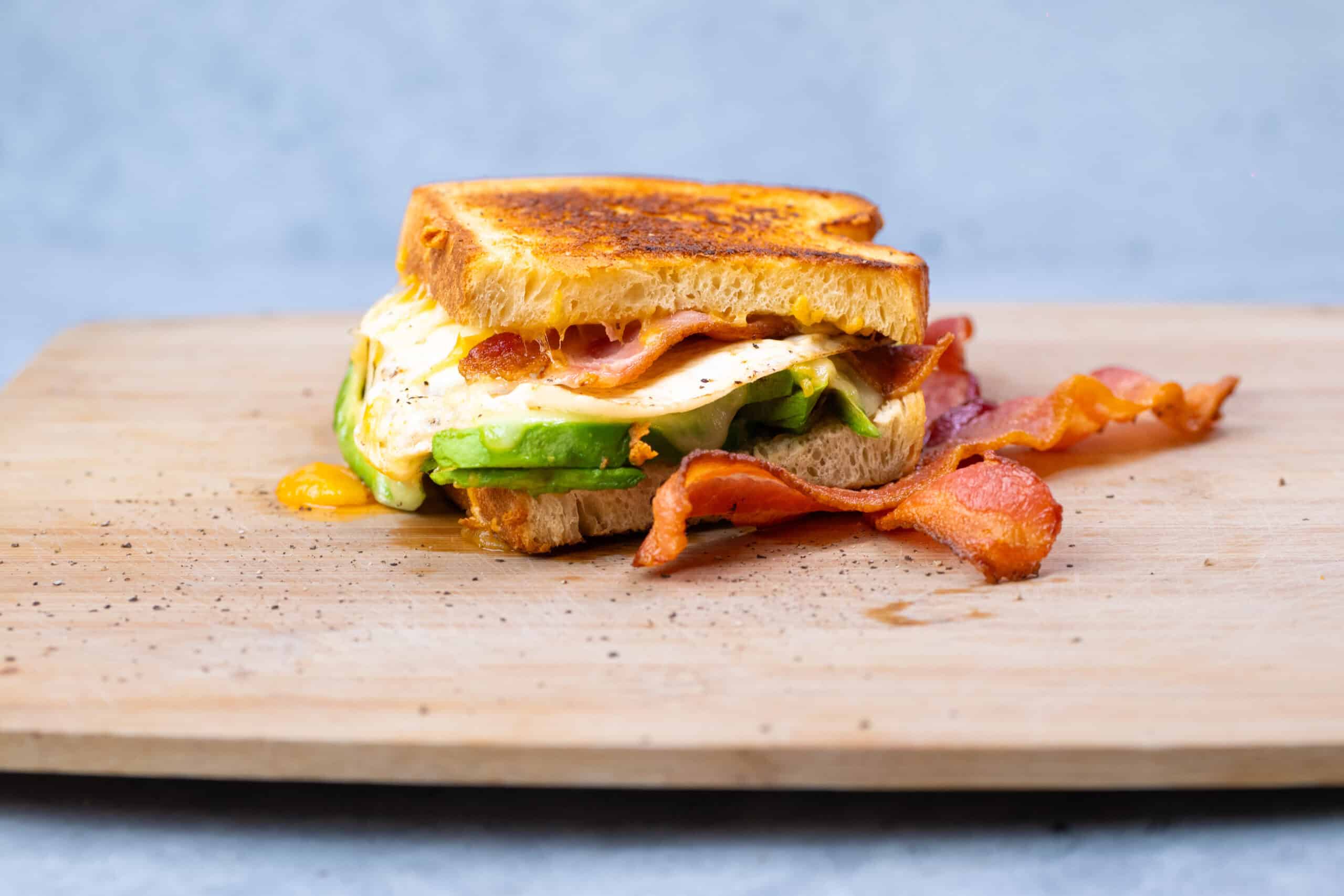 Savory breakfast sandwich with crispy bacon, fried egg, fresh avocado slices, and toasted bread on wooden cutting board. Perfect for morning meal or brunch.