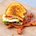 Savory breakfast sandwich with fried egg, crispy bacon, and fresh vegetables on toasted bread, served on a wooden cutting board.