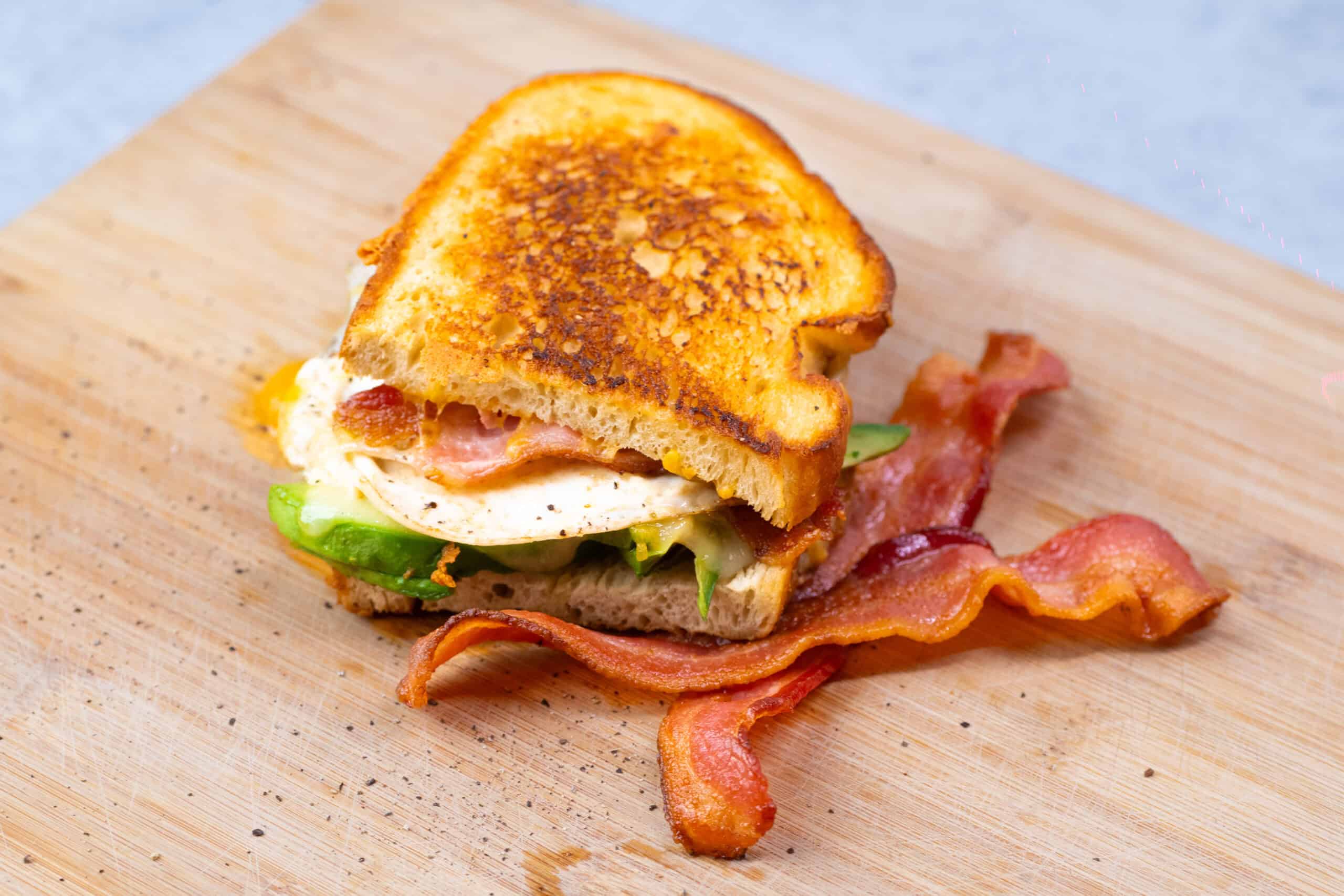 Savory breakfast sandwich with fried egg, crispy bacon, and fresh vegetables on toasted bread, served on a wooden cutting board.