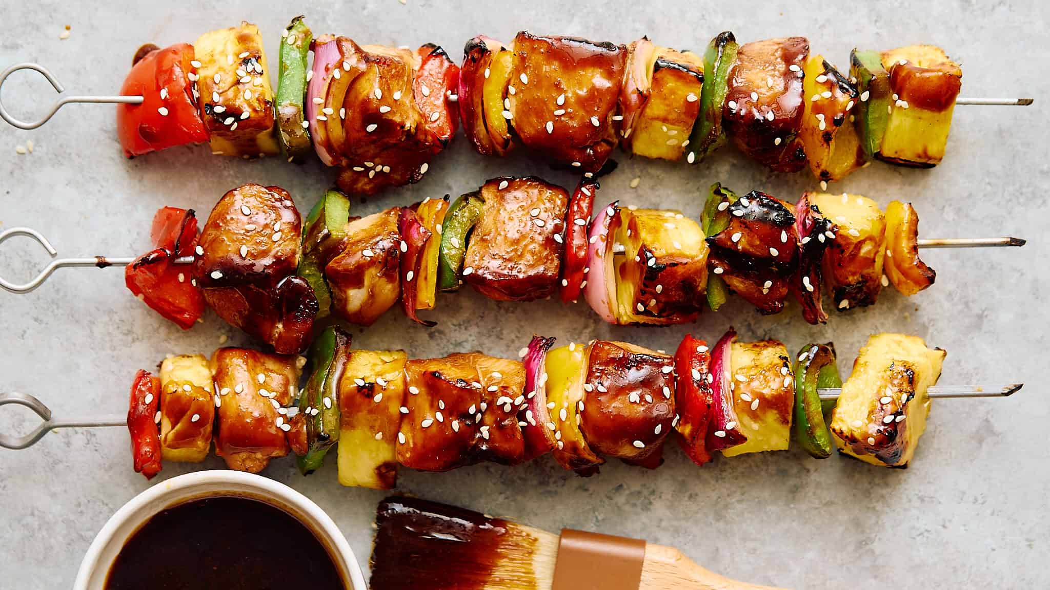 Colorful vegetable and chicken skewers drizzled with teriyaki sauce, garnished with sesame seeds, on a light gray surface.