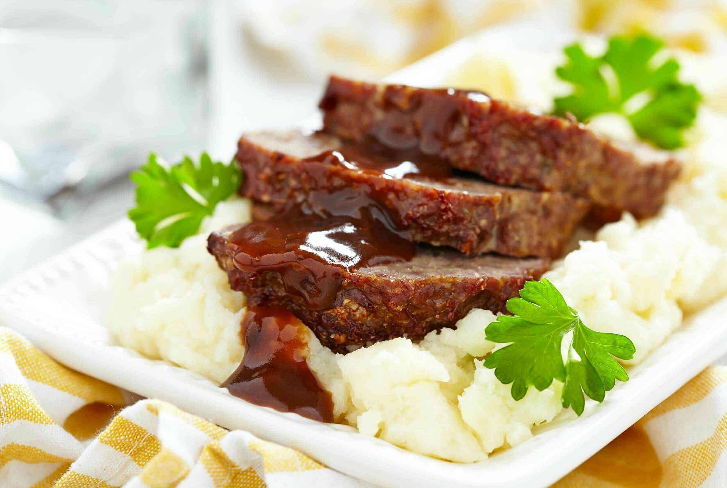 Succulent slices of meatloaf topped with rich brown gravy served over creamy mashed potatoes, garnished with fresh parsley on a white plate for a comforting home-cooked meal.
