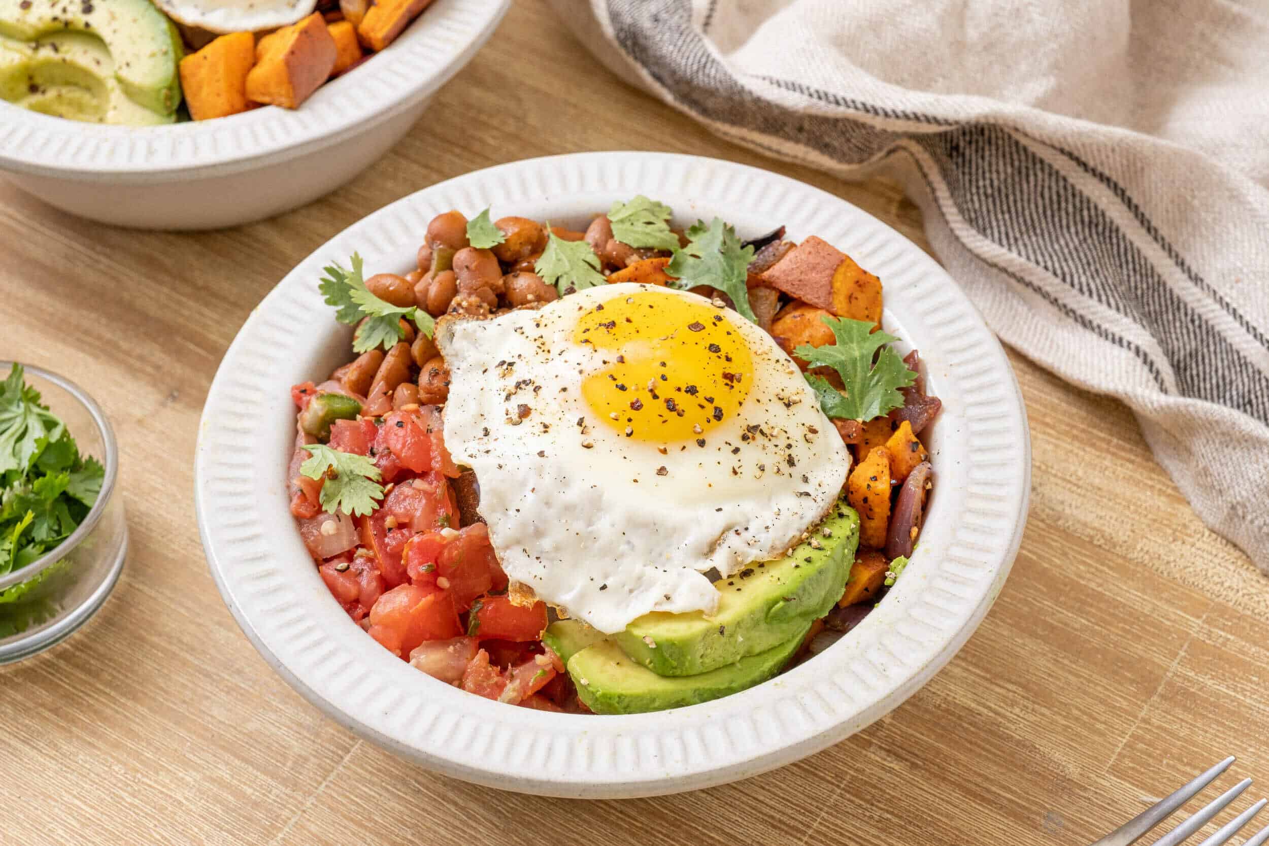 Fresh Mexican-style salad bowl with avocado, cooked beans, tomatoes, and a fried egg with black pepper on top, served in a white ceramic bowl on wooden table.