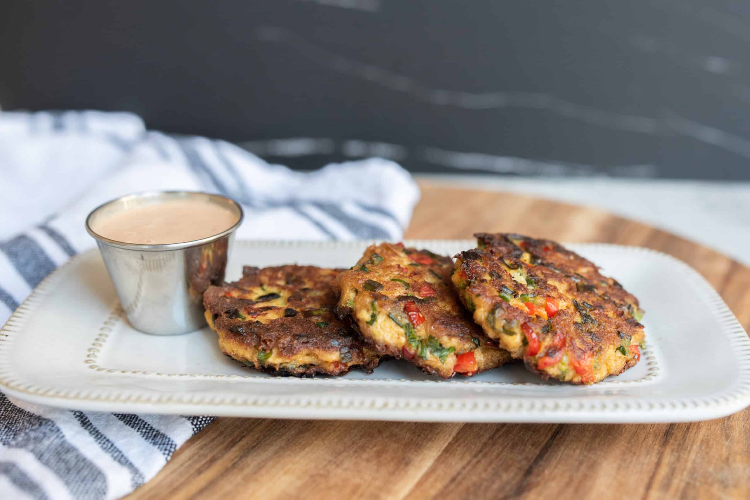 Crispy vegetable fritters served with a side of creamy dipping sauce on a white ceramic plate. Perfect for healthy snack or appetizer, packed with colorful chopped vegetables and herbs.