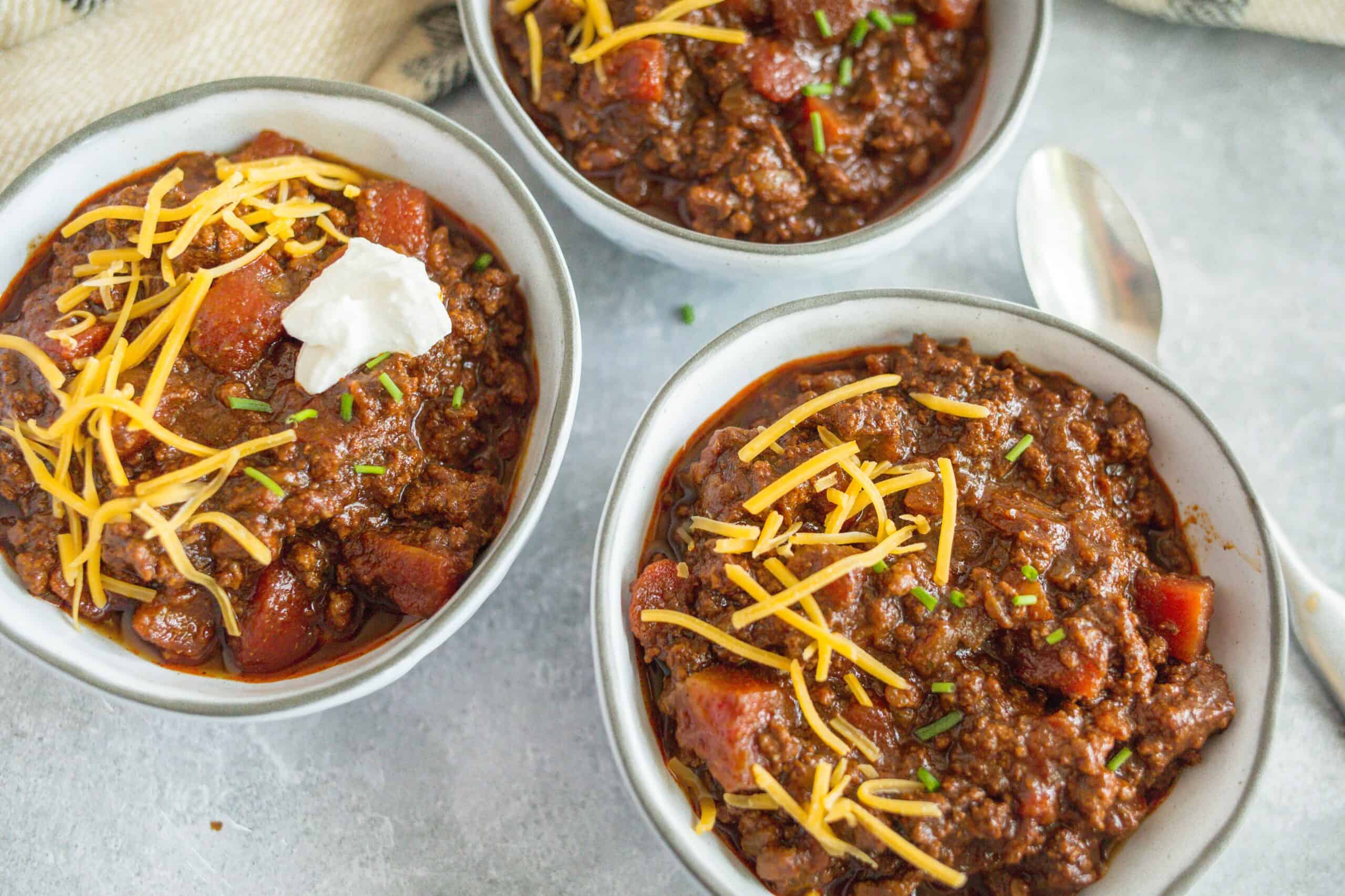 Chunky beef chili with tomatoes, cheese, and green onions in white bowls, served as a hearty comfort food.