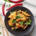 Succulent chicken stir-fry with basil and chili, served in a black bowl on a white rustic table, garnished with fresh herbs, accompanied by red chili peppers, chopsticks, and basil leaves for an Asian cuisine-inspired meal.