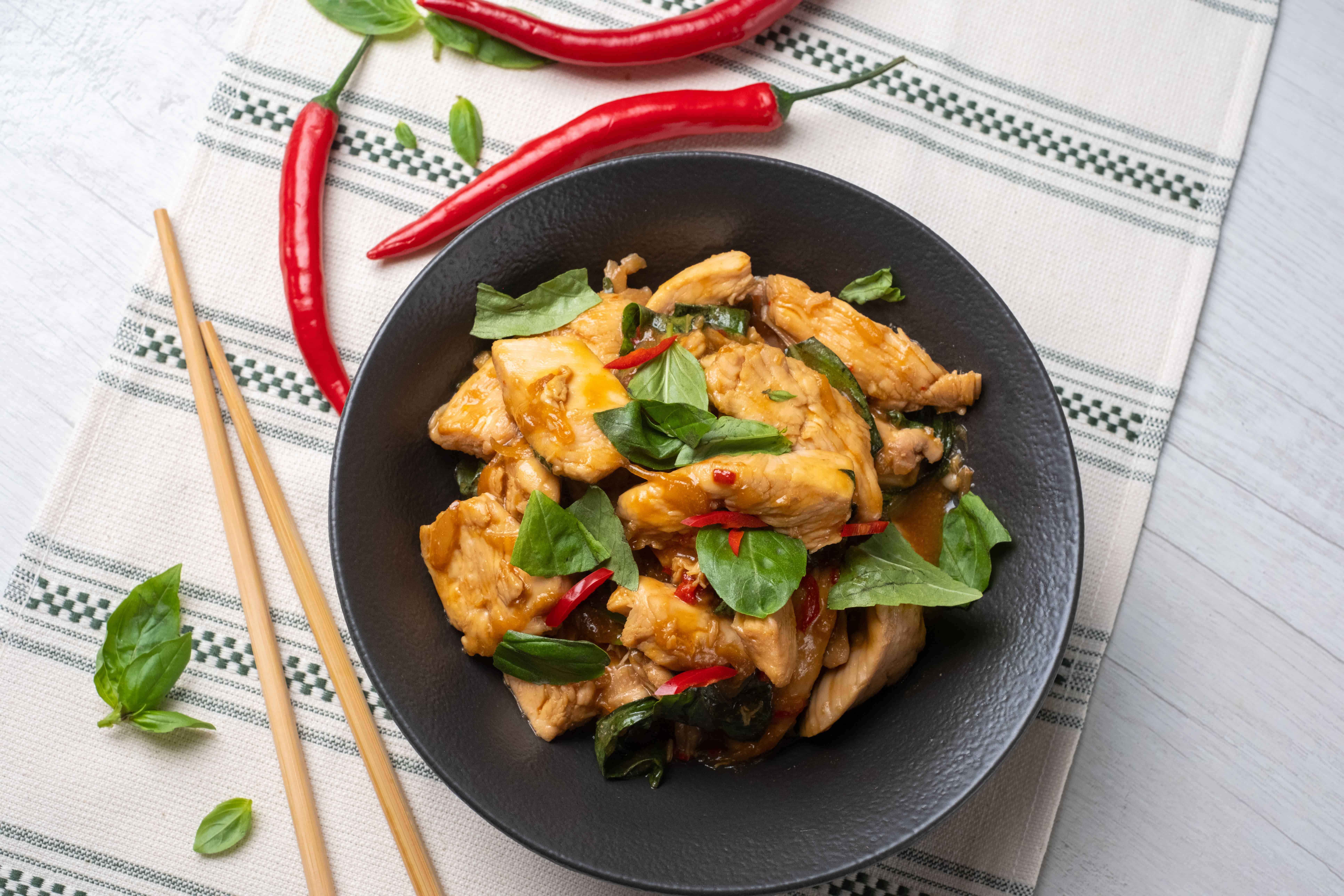 Succulent chicken stir-fry with basil and chili, served in a black bowl on a white rustic table, garnished with fresh herbs, accompanied by red chili peppers, chopsticks, and basil leaves for an Asian cuisine-inspired meal.