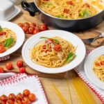 Spaghetti with cherry tomatoes and fresh basil on white plates, ingredients and uncooked pasta on wooden table, homemade Italian pasta dish, vibrant and delicious pasta presentation, perfect for Italian cuisine lovers.
