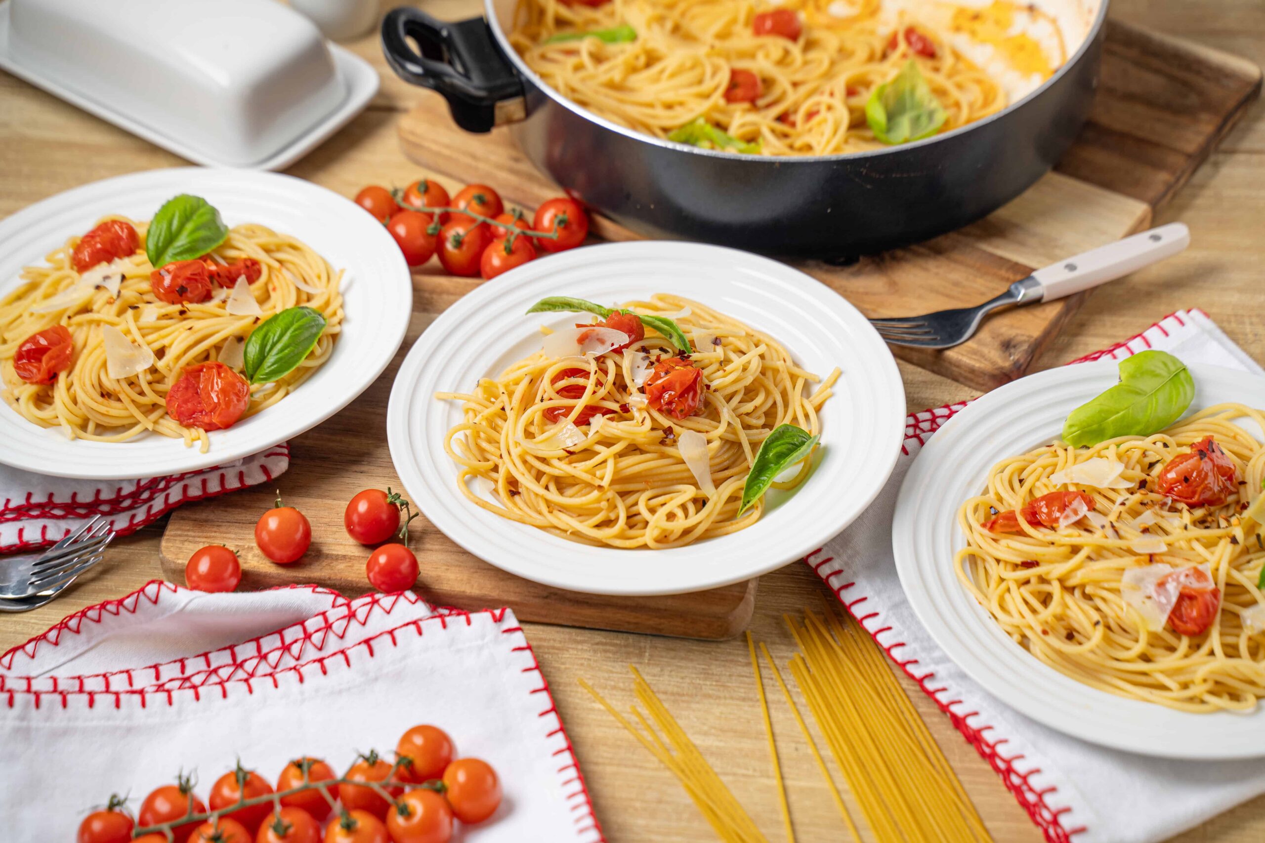 Spaghetti with cherry tomatoes and fresh basil on white plates, ingredients and uncooked pasta on wooden table, homemade Italian pasta dish, vibrant and delicious pasta presentation, perfect for Italian cuisine lovers.
