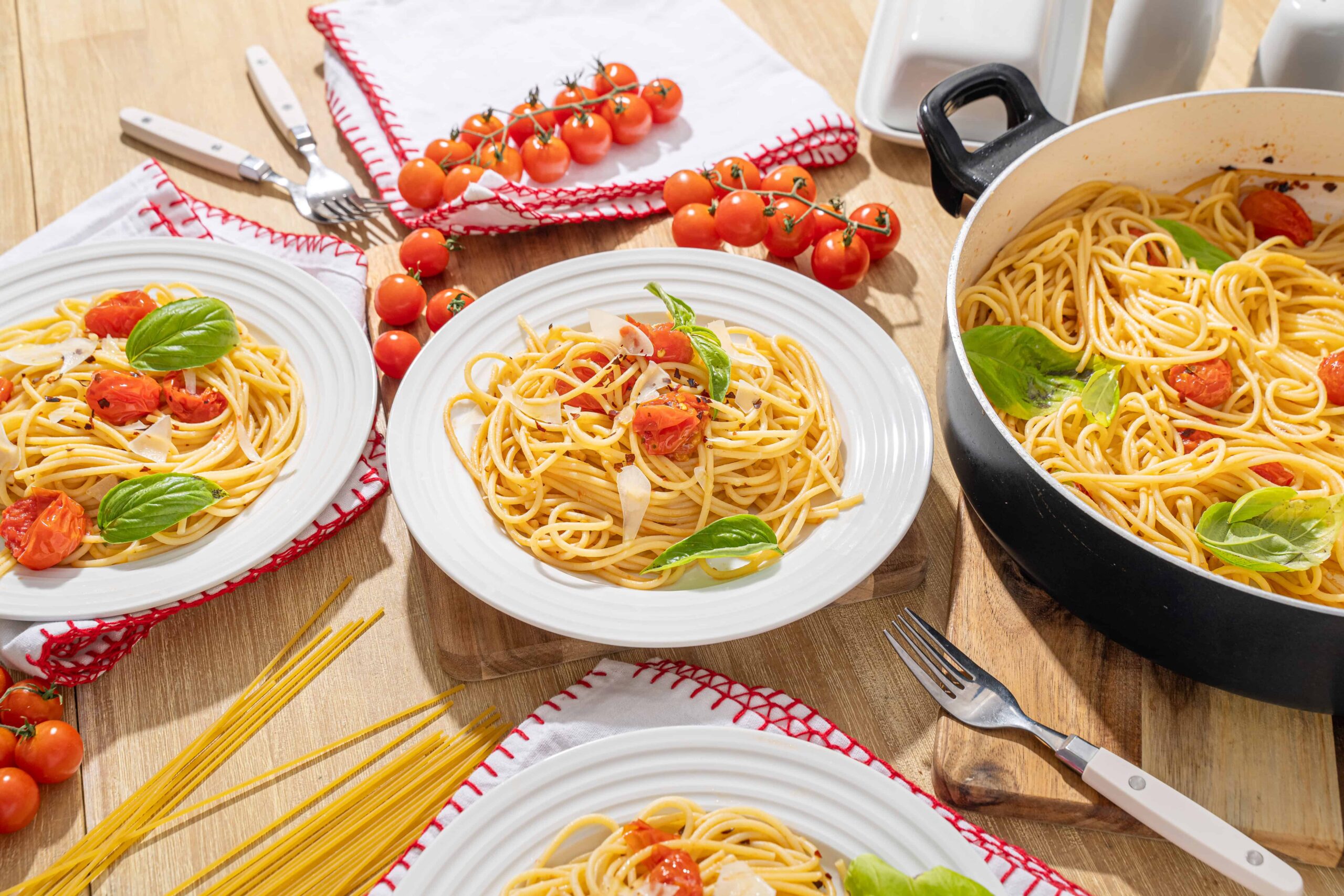 Vibrant spaghetti aglio e olio with cherry tomatoes, fresh basil, and Parmesan cheese served on white plates, with ingredients and kitchen essentials arranged on a wooden table.