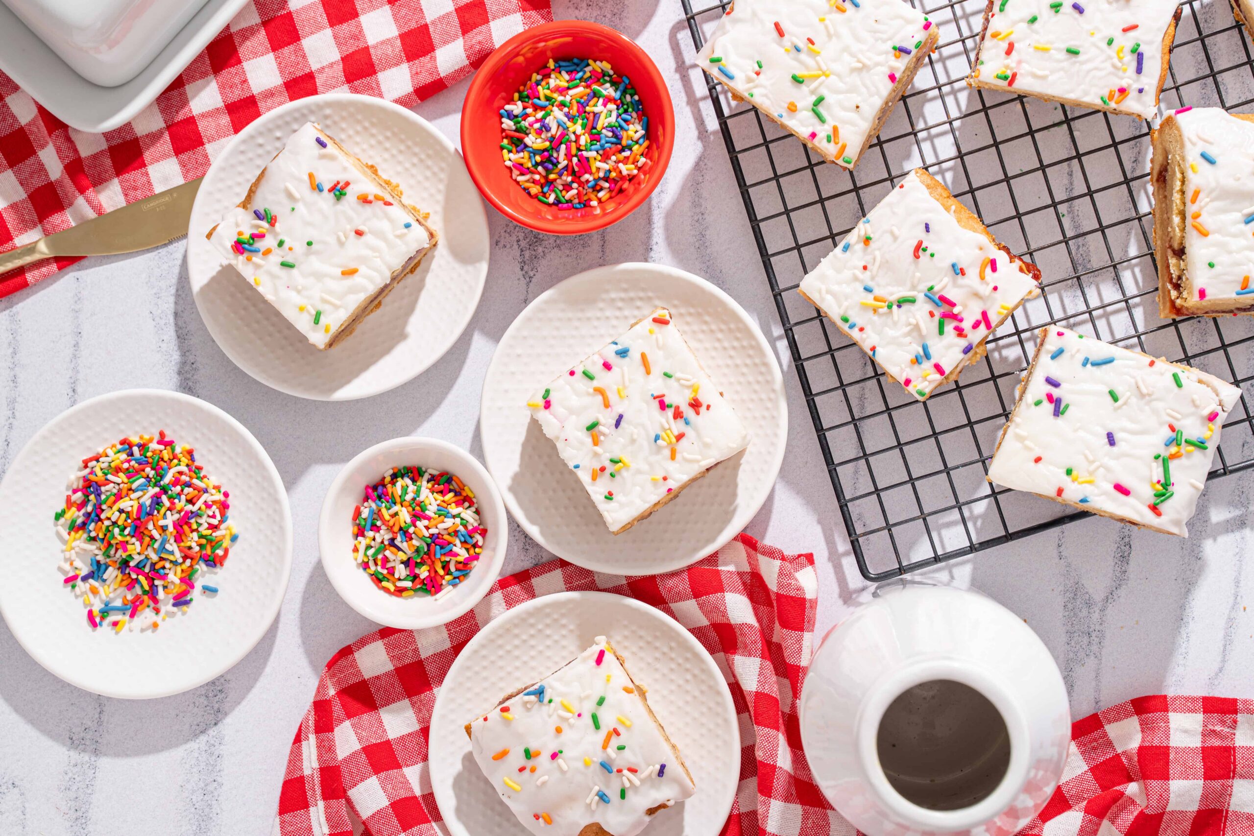 Brightly decorated homemade frosted sugar cookie bars topped with colorful rainbow sprinkles on white plates and a cooling rack, set on a white surface with red checkered cloths for a cheerful dessert presentation.
