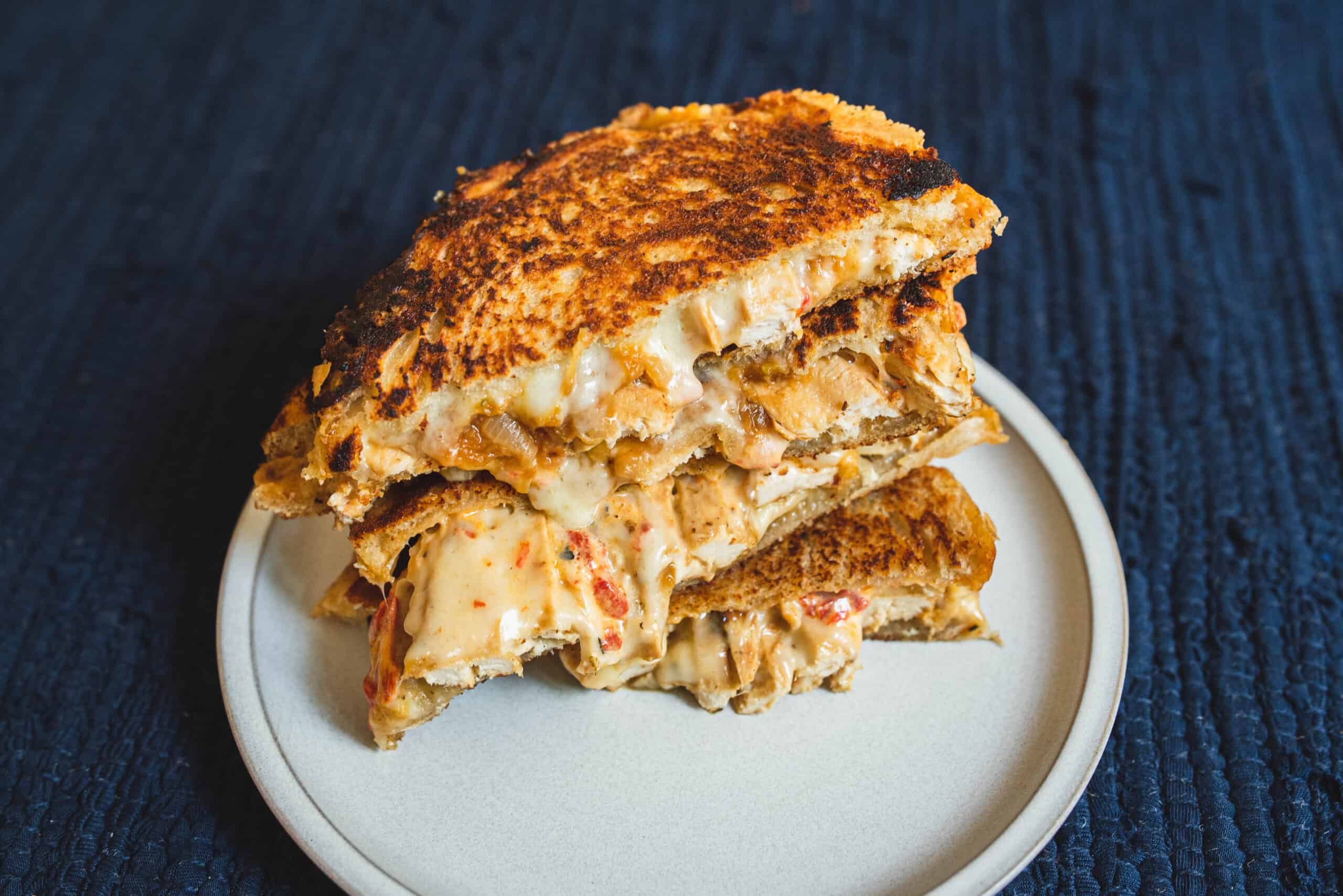 Crispy chicken cordon bleu sandwich with melted cheese on a white plate, close-up, dark blue textured background.