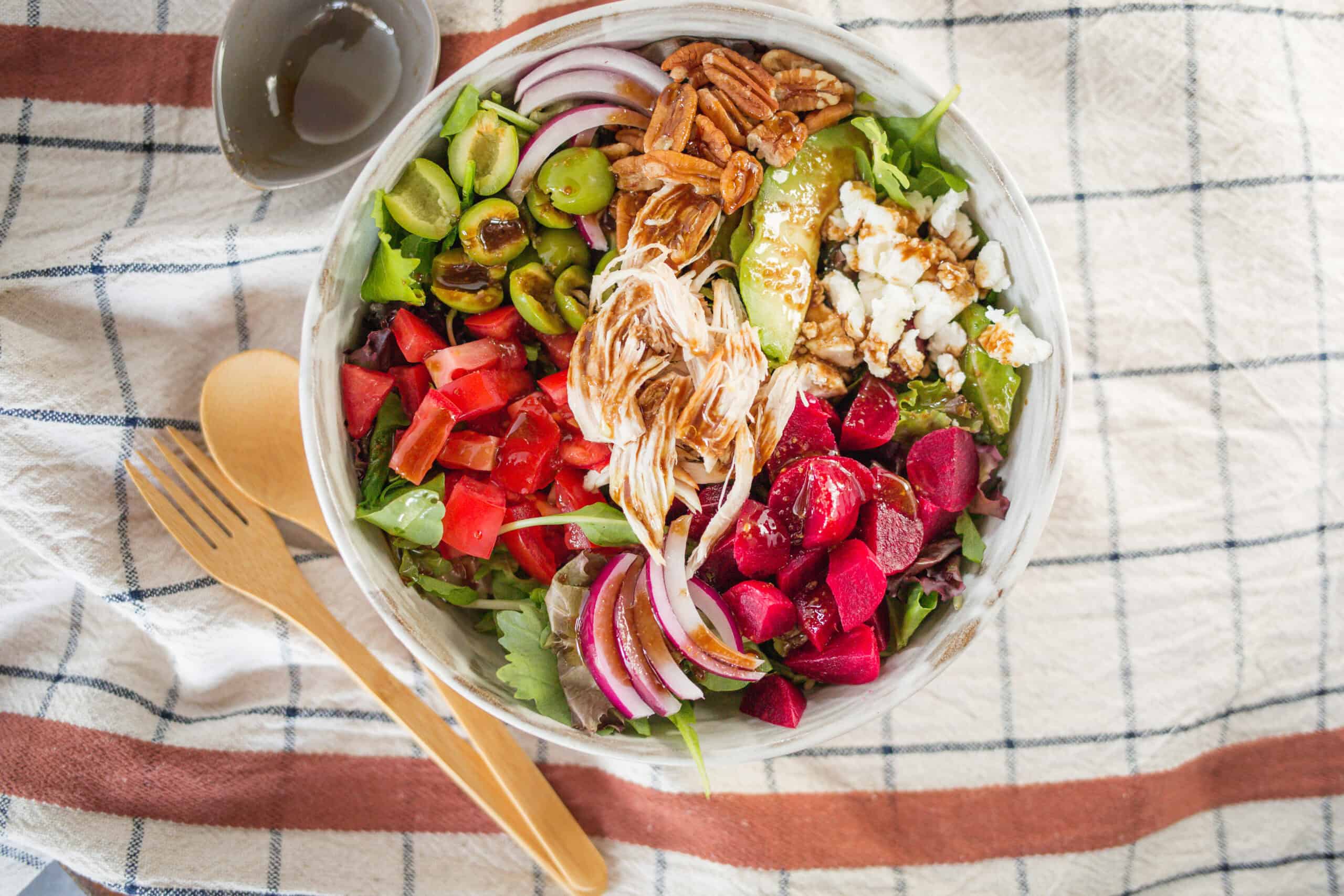 Fresh mixed vegetable salad with grilled chicken, nuts, and cheese, served in a white bowl on a checkered cloth. Includes ingredients like cherry tomatoes, radishes, cucumbers, red onions, alfalfa sprouts, and balsamic dressing.