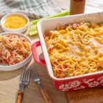 Creamy baked spaghetti casserole with melted cheese, served alongside fresh shredded cheese, cooked pasta, shredded chicken, and chopped vegetables for a hearty dinner option.