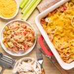 Baked chicken and vegetable spaghetti casserole topped with melted cheese in a baking dish surrounded by bowls of pasta, shredded chicken, celery sticks, and cheese on a wooden table.