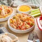 Creamy chicken spaghetti casserole with shredded chicken, pasta, and vegetables, served in white bowls on a rustic wooden table. Includes shredded cheese, celery sticks, and side dishes.