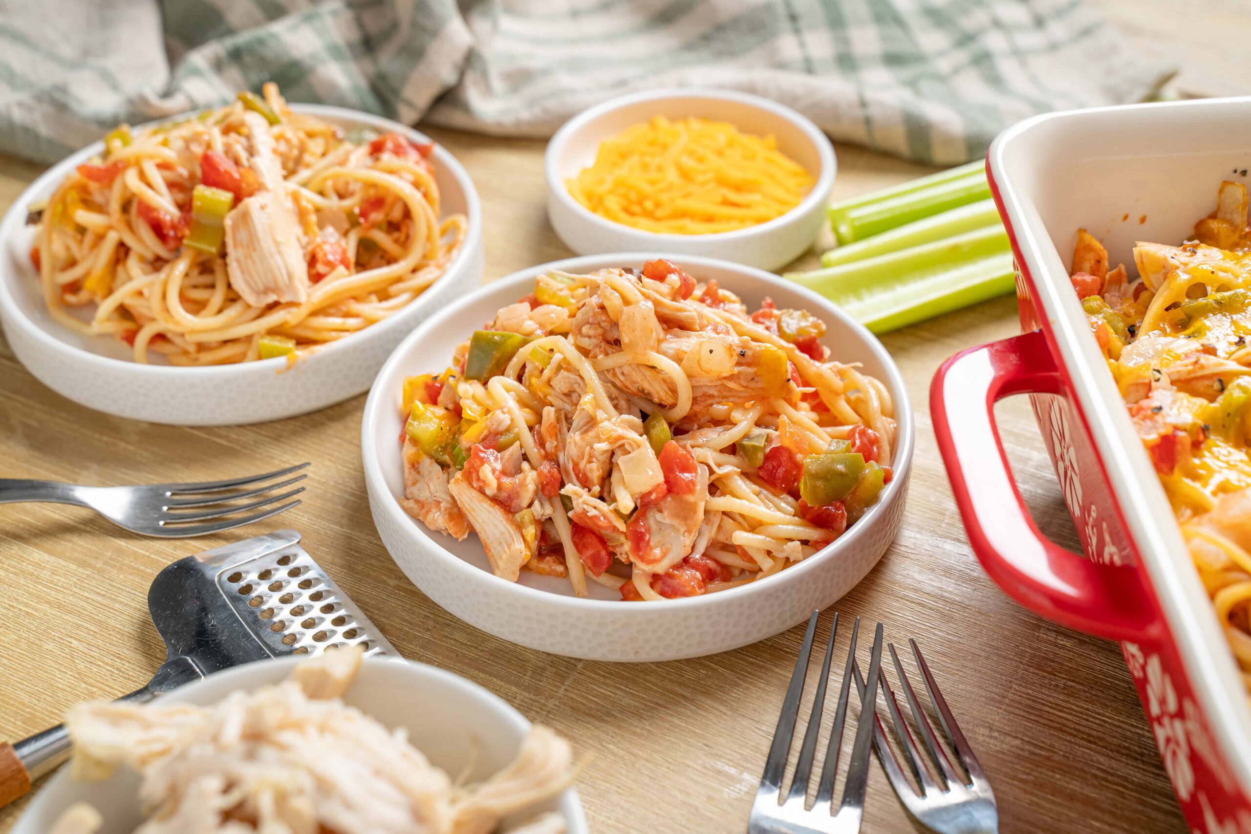 Creamy chicken spaghetti casserole with shredded chicken, pasta, and vegetables, served in white bowls on a rustic wooden table. Includes shredded cheese, celery sticks, and side dishes.