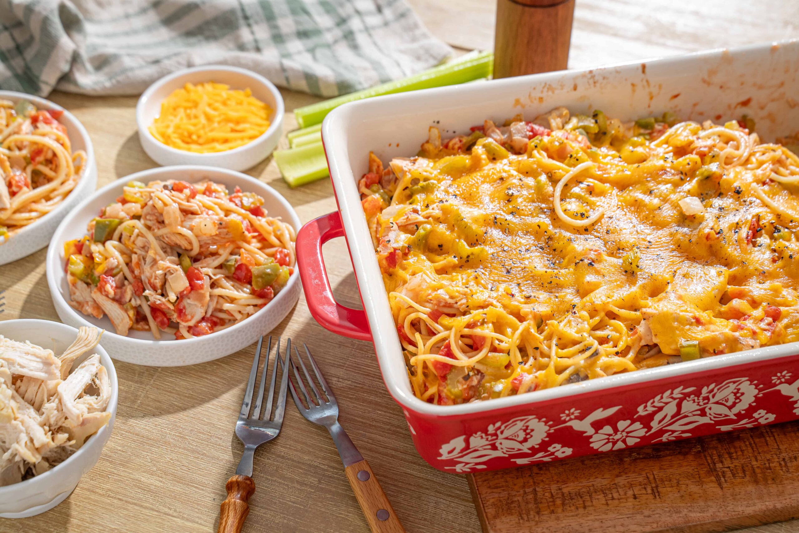 Creamy baked spaghetti casserole with melted cheese, served alongside fresh shredded cheese, cooked pasta, shredded chicken, and chopped vegetables for a hearty dinner option.